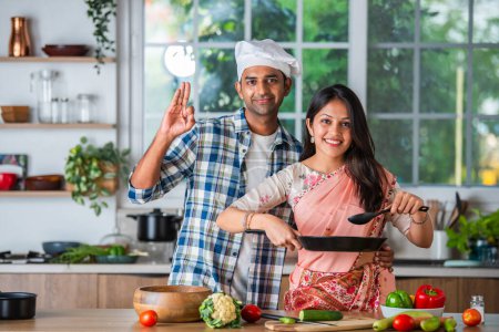 Photo for Indian asian married couple in kitchen promoting utensils, empty dish with ok sign - Royalty Free Image