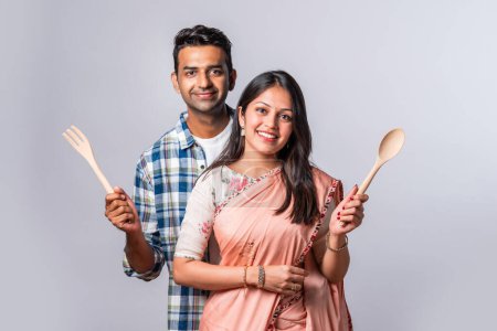 Photo for Portrait of smiling Indian asian young couple with wooden and metal kitchen utensils, standing isolated on white - Royalty Free Image