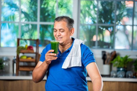 Téléchargez les photos : Indien asiatique beau moyen âge l'homme boire sain vert boisson ou smoothie dans cuisine - en image libre de droit