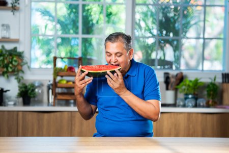 Téléchargez les photos : Indian asian mid age man enjoy eating watermelon fruit over white or kitchen background - en image libre de droit