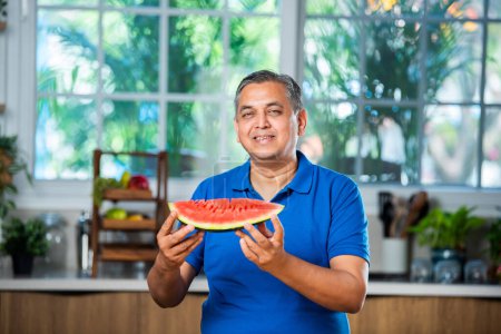 Téléchargez les photos : Indien asiatique moyen âge homme profiter manger pastèque fruits sur fond blanc ou cuisine - en image libre de droit