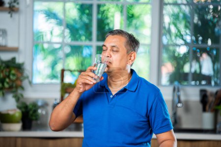 Foto de Indio asiático mediana edad guapo hombre bebiendo llano agua en un vaso en casa - Imagen libre de derechos