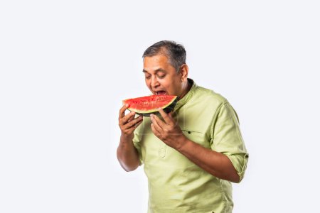Téléchargez les photos : Indien asiatique moyen âge homme profiter manger pastèque fruits sur fond blanc ou cuisine - en image libre de droit