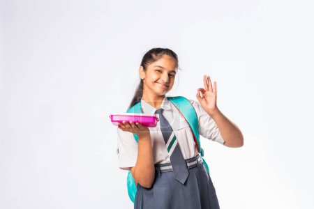 Photo for Happy Indian asian schoolgirl in school uniform holding a lunch box against white background - Royalty Free Image