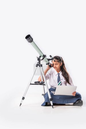 Téléchargez les photos : Petite écolière indienne asiatique en uniforme scolaire avec télescope sur fond blanc - en image libre de droit