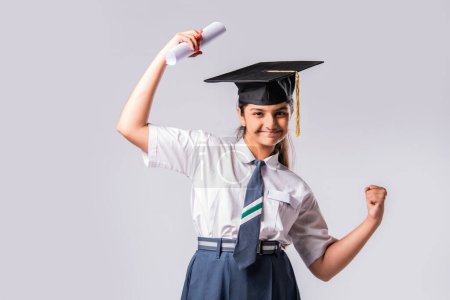 Photo for Happy Asian Indian schoolgirl wears school uniform and graduation cap against white background - Royalty Free Image