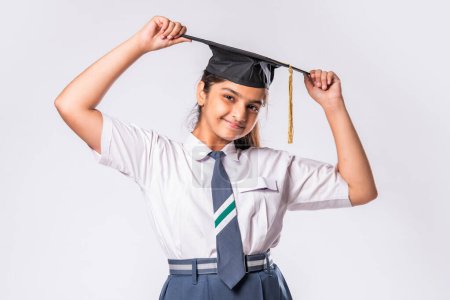 Téléchargez les photos : Heureuse écolière indienne asiatique porte uniforme scolaire et chapeau de graduation sur fond blanc - en image libre de droit