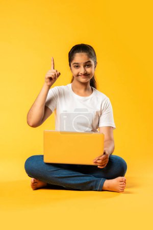 Photo for Indian asian pretty small school girl using laptop sitting isolated on yellow background - Royalty Free Image