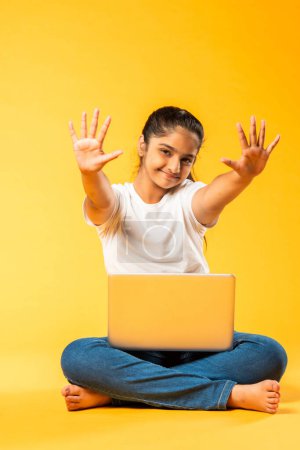 Photo for Indian asian pretty small school girl using laptop sitting isolated on yellow background - Royalty Free Image