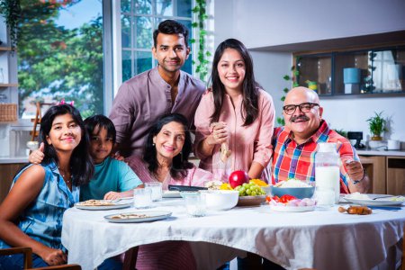 Foto de Retrato de la familia india de tres generaciones comiendo juntos en casa mirando a la cámara - Imagen libre de derechos