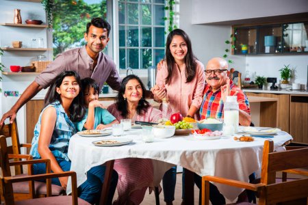 Foto de Retrato de la familia india de tres generaciones comiendo juntos en casa mirando a la cámara - Imagen libre de derechos