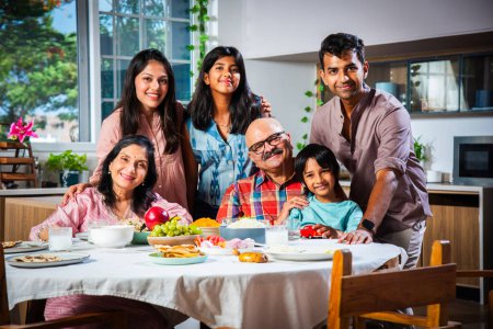 Téléchargez les photos : Portrait de famille indienne de trois générations mangeant ensemble à la maison en regardant la caméra - en image libre de droit