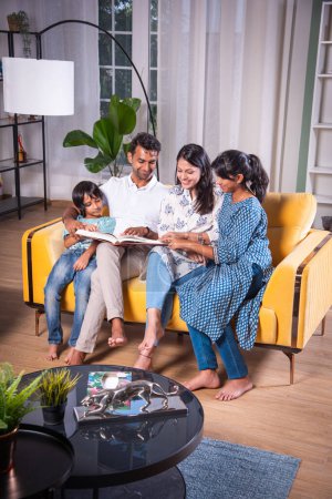 Foto de India asiática joven familia de cuatro mirando fotos en álbum de fotos o libro de lectura juntos sentado en el sofá - Imagen libre de derechos