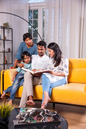 Photo for Indian asian young family of four looking at photos in photo album or reading book together sitting on sofa - Royalty Free Image