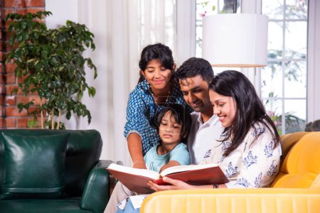Photo for Indian asian young family of four looking at photos in photo album or reading book together sitting on sofa - Royalty Free Image