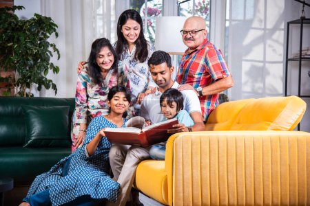 Photo for Indian asian multigenerational family watching photos in album or reading book while sitting on sofa together - Royalty Free Image
