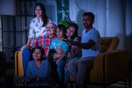 Photo for Happy Indian asian multigenerational family watching television or tv while sitting on sofa at home - Royalty Free Image
