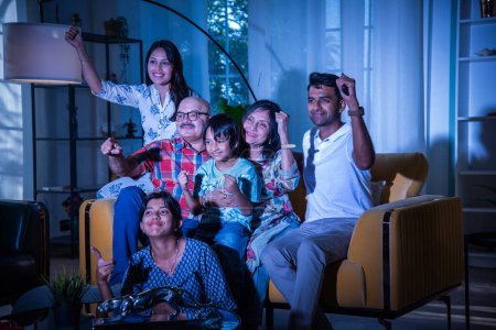 Happy Indian asian multigenerational family watching television or tv while sitting on sofa at home