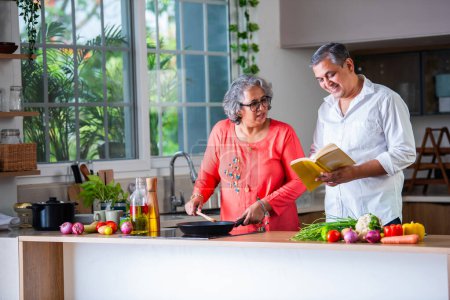 Indian asian senior retired old couple reading recipe book while cooking in the kitchen together