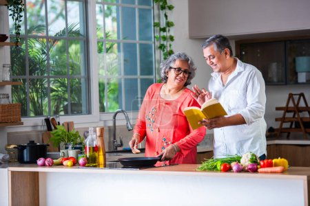 Indian asian senior retired old couple reading recipe book while cooking in the kitchen together