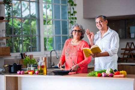 Téléchargez les photos : Indien asiatique senior retraité vieux couple lecture recette livre tandis que cuisson dans l 'cuisine ensemble - en image libre de droit