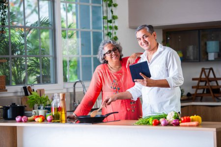 Téléchargez les photos : Indien asiatique couple aîné en utilisant ordinateur portable ou tablette dans la cuisine pour la commande ou les médias sociaux - en image libre de droit