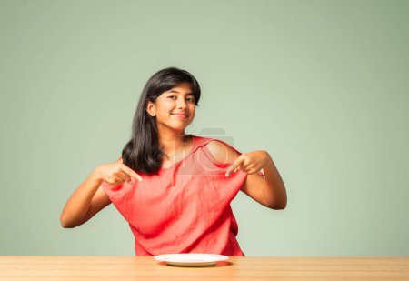 Foto de Indio asiático pequeña chica presentando vacío plato mientras sentado en mesa - Imagen libre de derechos