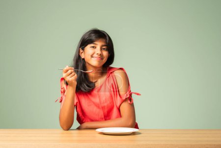 Photo for Indian asian small girl presenting empty plate while sitting at table - Royalty Free Image