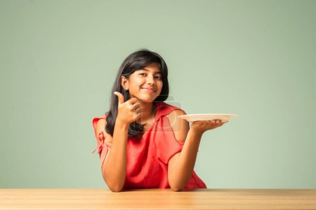 Photo for Indian asian small girl presenting empty plate while sitting at table - Royalty Free Image