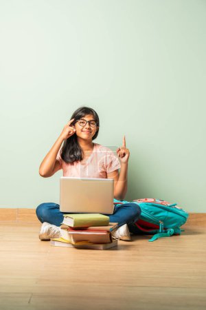 Téléchargez les photos : Indien asiatique école fille en casual porter à l'aide d'ordinateur portable pour étude assis sur sol isolé - en image libre de droit