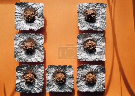 Foto de Siete pralinés de chocolate esférico se despliegan en envolturas de papel de aluminio. Fondo naranja, espacio para copiar. Vista superior, rectángulo, cuadrado. - Imagen libre de derechos