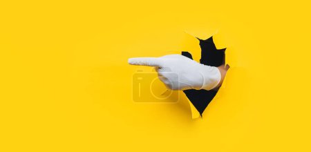 Photo for The forefinger points in white medical glove to the left side. Yellow paper background with torn hole. Place for advertising. Copy space. - Royalty Free Image