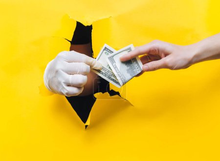 Photo for Hand of a woman patient gives money in the form of hundred-dollar bills to the doctor in a white medical glove. Yellow paper background, torn hole. The concept of payment for treatment. Gynecology. - Royalty Free Image