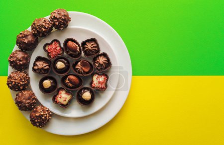 Photo for Beautiful chocolates of different shapes and fillings lie in a white plate against the yellow and green background paper. Copy space, top view. - Royalty Free Image