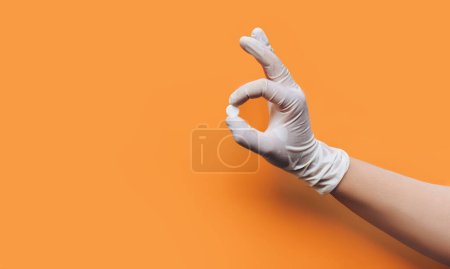 Téléchargez les photos : La main d'un médecin dans un gant de caoutchouc médical blanc tient une tablette (pilule) avec deux doigts sous la forme d'un symbole OK. Fond orange, espace de copie. Concept. - en image libre de droit