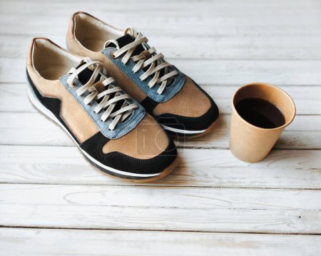 Foto de Un par de zapatillas de gamuza beige sobre un fondo grunge de madera y una taza de papel de café. El concepto de bebida energética, calzado deportivo y sed. - Imagen libre de derechos