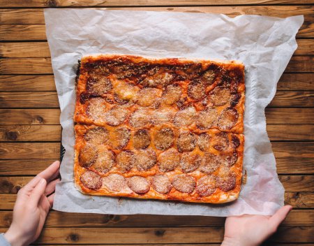 Foto de Las manos de las mujeres sostienen forma de rectángulo de pizza casera con queso y salami en papiro sobre el fondo de una mesa de madera marrón. Vista desde arriba. - Imagen libre de derechos