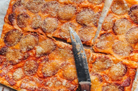 Téléchargez les photos : Forme rectangle de pizza maison fraîche avec du fromage et du salami se trouve sur le papyrus sur le fond d'une table en bois brun fermer. Couteau pour couper. Vue d'en haut, gros plan. - en image libre de droit