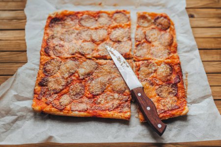 Foto de Deliciosa forma de rectángulo de pizza casera con queso y salami se encuentra sobre el papiro en el fondo de una mesa de madera marrón de cerca. Vista desde arriba. Cuchillo para cortar. - Imagen libre de derechos