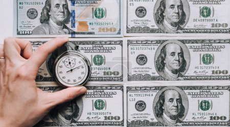 Téléchargez les photos : C'est l'heure de l'argent. Main de femme tenant un chronomètre gris sur fond de dollars. Concept de crédit. - en image libre de droit