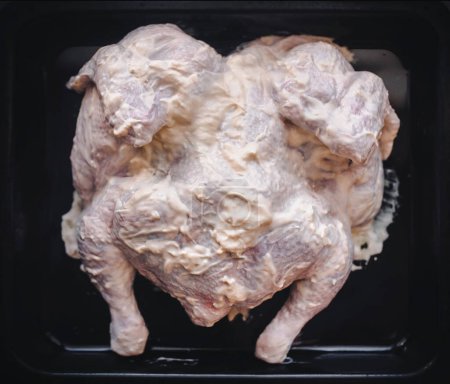 Photo for Raw chicken is covered with seasonings in a mayonnaise marinade. The recipe for baking chicken in the oven. Black dripping pan, top view. - Royalty Free Image