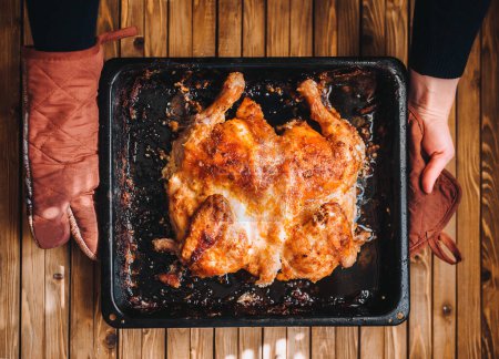 Photo for A pan with roast turkey in female hands. Thanksgiving Day. Homemade roasted chicken in a black pan on a wooden background. Lifestyle. Top view. - Royalty Free Image