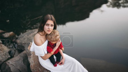 Téléchargez les photos : Jeune femme mannequin avec un visage triste assis sur les rochers près de la rivière avec une poupée sur ses genoux. Une adolescente imagine qu'elle est une mère. - en image libre de droit