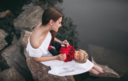 Photo for Young model-looking woman sitting on the rocks near the river with a doll on her lap. A teenage girl imagines that she is a mother. - Royalty Free Image