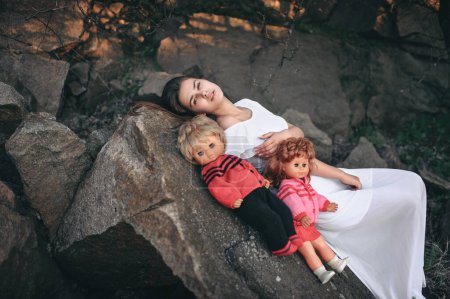 Téléchargez les photos : Jeune femme modèle se trouve sur les rochers avec deux poupées. Une adolescente imagine qu'elle est une mère. - en image libre de droit