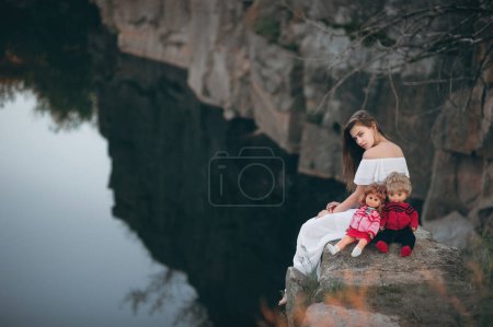 Photo for Young model-looking woman sitting on the rocks near the river with two dolls. A teenage girl imagines that she is a mother. - Royalty Free Image