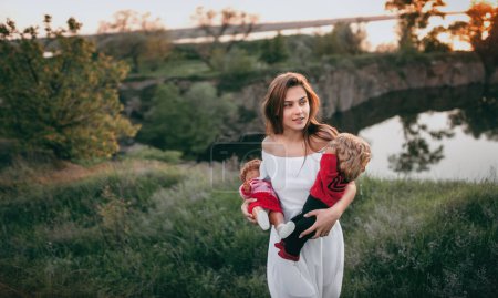 Photo for Young model-looking woman walking along the field near the river with two dolls in her hands. A teenage girl imagines that she is a mother. - Royalty Free Image