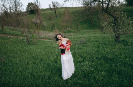 Foto de Mujer joven de aspecto modelo con una cara feliz caminando por el campo cerca del río con dos muñecas en sus manos. Una adolescente se imagina que es madre. - Imagen libre de derechos