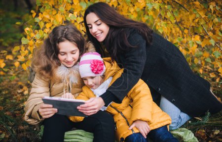 Téléchargez les photos : Une jeune mère et ses filles regardent quelque chose d'intéressant sur la tablette tout en marchant dans le parc. - en image libre de droit