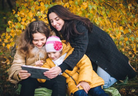 Téléchargez les photos : Une jeune mère et ses filles regardent quelque chose d'intéressant sur la tablette et sourient en marchant dans le parc. - en image libre de droit